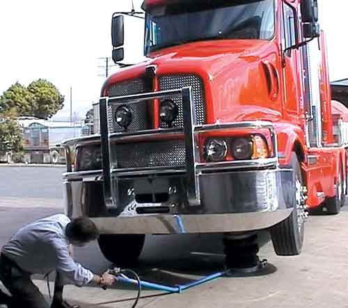 A man operating a heavy duty Selson Air Jack to lift up the front left corner of a red haulage truck.